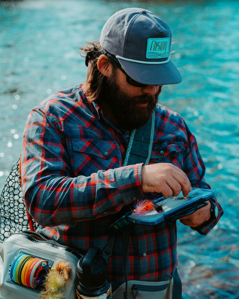 Man fishing by a river, wearing a cap and plaid jacket.