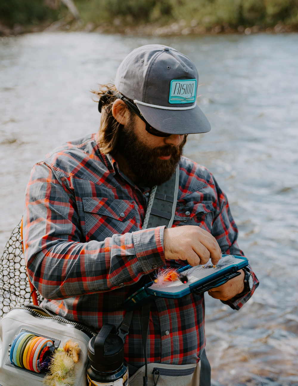 Man fishing by a river, wearing a cap and plaid jacket.